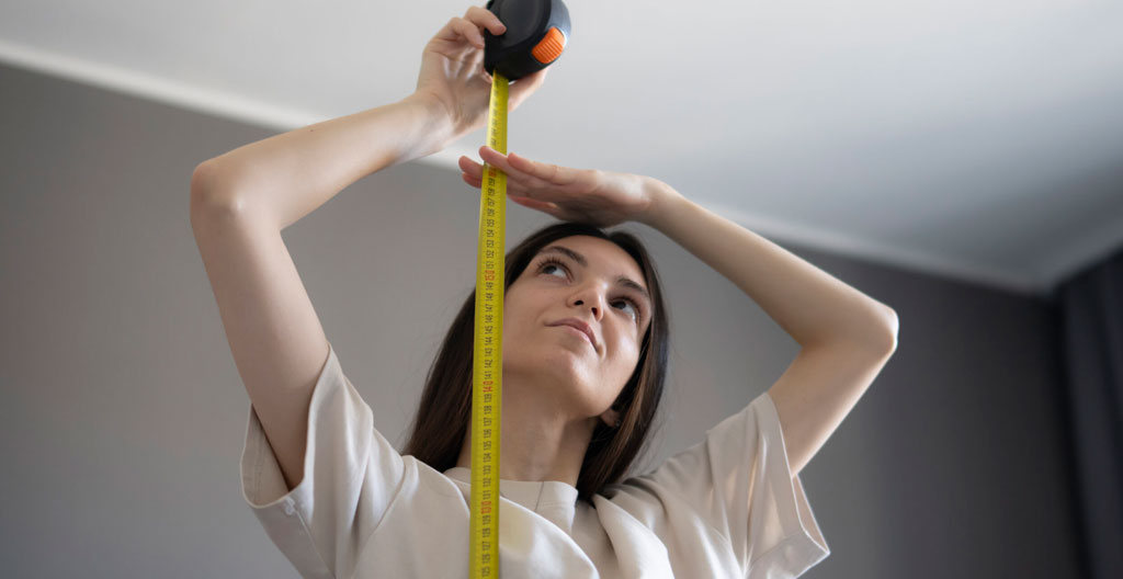 Femme trentenaire en jean et pull beige mesurant hauteur sous plafond avec télémètre laser rouge dans pièce vide aux murs blancs et parquet chêne clair, lumière naturelle douce